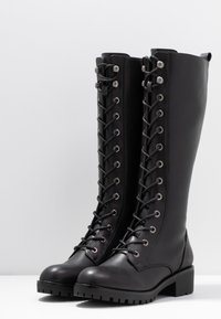 Black leather knee-high boots with a lace-up front, silver eyelets, and a chunky sole. Smooth texture and flat block heel.
