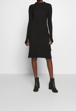 Frau trägt ein schwarzes, geripptes Midi-Kleid mit langen Ärmeln, seitlichen Schlitzen und schwarzen Stiefeletten, steht vor einem weißen Hintergrund.