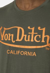 Primer plano de una camiseta verde con texto naranja desvanecido que dice "Von Dutch" y "CALIFORNIA" en el pecho.