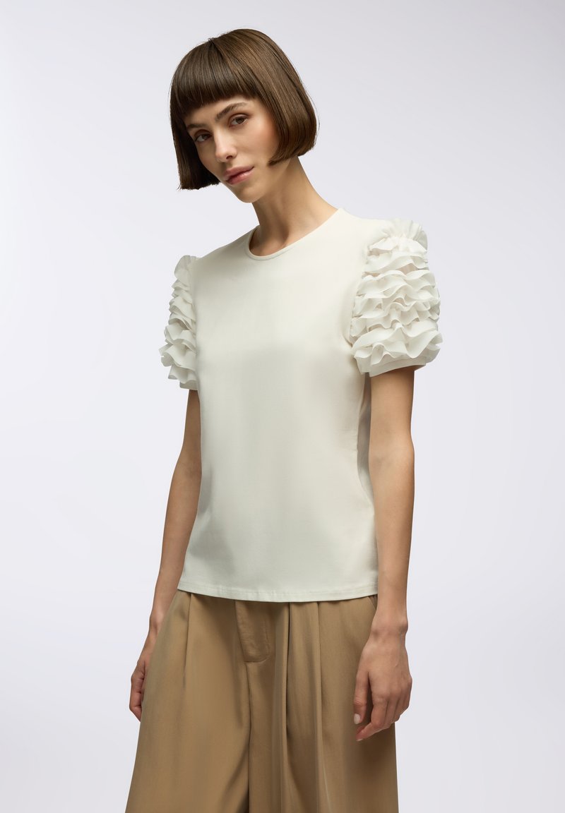 Top bianco con maniche corte a volants, scollatura rotonda e texture liscia. Abbinato a pantaloni beige larghi, senza pattern o hardware visibili.