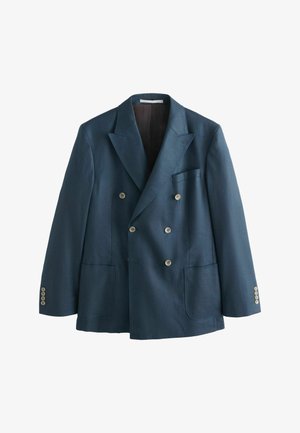 Mężczyzna w granatowym garniturze dwurzędowym, białej koszuli zapinanej na guziki oraz wzorzystym brązowym krawacie na neutralnym tle.