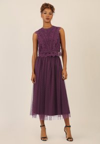 Violetti midi-mekko, jossa on pitsinen yläosa, jossa on aaltoilevat reunat, verkkomateriaali, jossa on pilkkuja, ja mustat korkokengät.