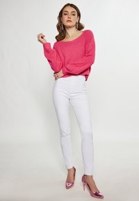 Maglione rosa fuzzy con scollatura ampia, jeans skinny bianchi e scarpe con tacco alto rosa lucido. Modella in posa su uno sfondo neutro.