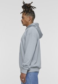 Light gray hoodie with a front pocket, oversized fit, ribbed cuffs and hem, worn over blue jeans. Hoodie features a drawstring neck.