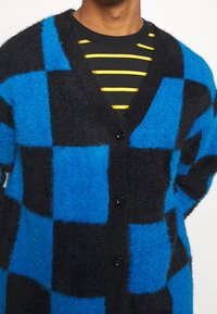 Untuvainen kardigan, jossa on sininen ja musta ruutukuvio, V-pääntie ja viisi mustaa nappia, päälle puettuna musta ja keltainen raitapaita.