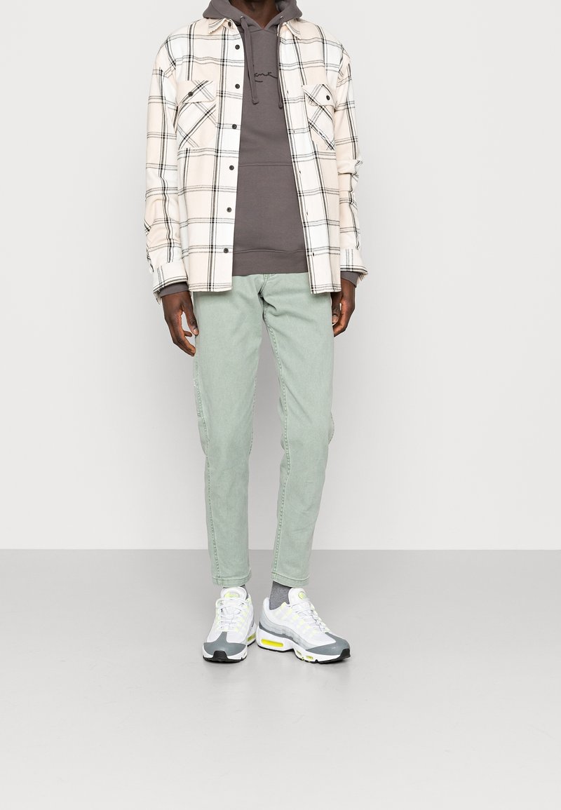 Homme portant un sweat à capuche gris sous une chemise à carreaux crème, un pantalon vert clair, des chaussettes grises et des baskets blanches avec des touches jaunes.