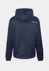 Námořnická bunda s kapucí vyrobená z lehkého materiálu s elastickými manžetami, na horní části zad s bílým logem "HILFIGER". Hladká textura.