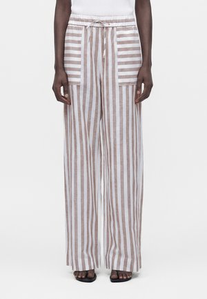 Femme portant un pantalon taille haute à jambes larges avec des rayures verticales beige et blanches et des poches plaquées, associé à des sandales noires et un haut blanc.