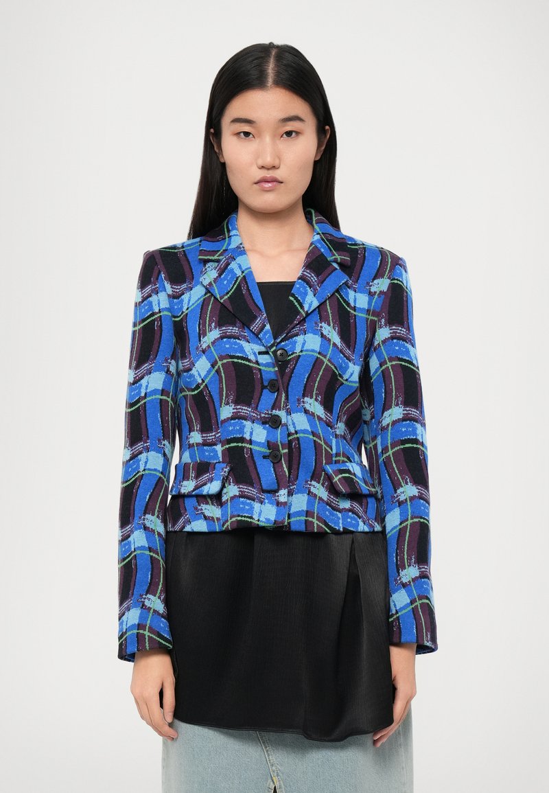 Blazer blu e nero a fantasia con colletto a punta, due tasche frontali e chiusura a bottone. Indossato sopra un top nero e una gonna di jeans.