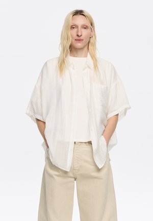 Camicia bianca a maniche corte con texture e righe sottili, abbinata a una maglietta bianca semplice e pantaloni a gamba larga beige chiaro.