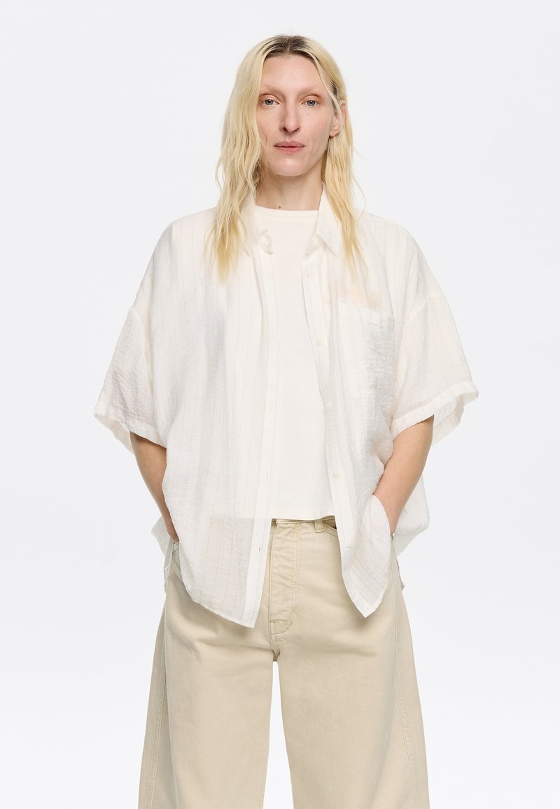 Camicia bianca a maniche corte con texture e righe sottili, abbinata a una maglietta bianca semplice e pantaloni a gamba larga beige chiaro.