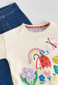 Tricou din bumbac crem cu broderii colorate, inclusiv un gândăcel, flori și fluturi. Asortat cu pantaloni din denim albastru închis.