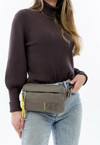 Šedá látková ledvinka s texturovaným povrchem, která má žlutý akcent a zipové uzavření. Nosená na pase přes džíny.