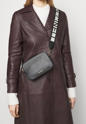 Czarna skórzana torebka na ramię z fakturowanym wykończeniem, złotym logo i szerokim, wzorzystym paskiem w czarno-białe wzory, noszona na bordowym płaszczu.