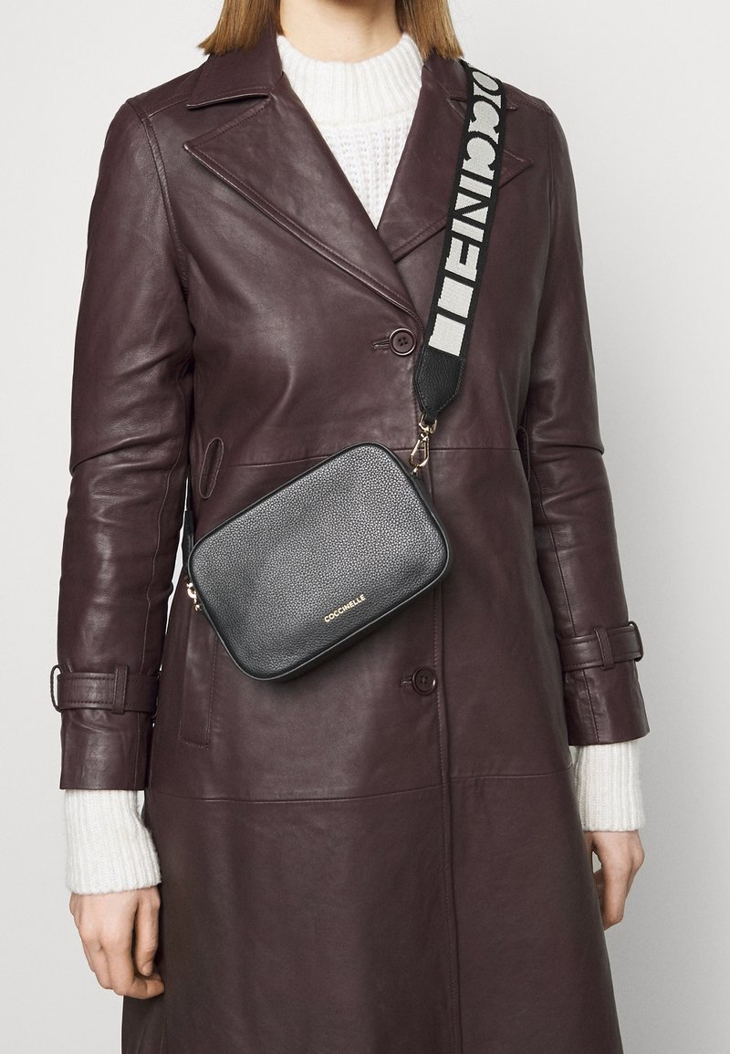 Černá kožená kabelka přes rameno s texturovaným povrchem, zlatým logem a širokým černobílým vzorovaným popruhem, nošená přes vínový kabát.