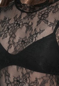 Černý květinový krajkový top navrstvený přes černou podprsenku, odhalující složitý průsvitný krajkový vzor a výstřih, s krátkými hnědými vlasy nahoře.