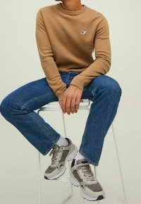 Beige neulepaita pyöreällä kaula-aukolla, vaaleansiniset farkut ja harmaat lenkkarit korostuksilla. Istuu valkoisella jakkaralla neutraalin taustan edessä.