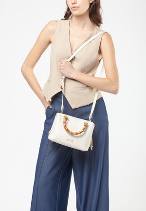 Witte leren handtas met een bamboe handvat, voorzien van een ritssluiting en reliëfdetails. Het model draagt een beige vest en blauwe broek.