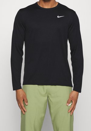 Homme portant un t-shirt noir à manches longues avec un logo Nike blanc sur la poitrine et un pantalon vert clair avec un petit logo blanc sur la cuisse.