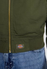 Veste bomber vert olive en tissu texturé, avec une fermeture éclair dorée et une taille côtelée, ornée d'un patch rectangulaire au logo Dickies.