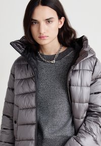 Veste matelassée grise avec capuche, comportant des sections matelassées et une fermeture éclair à l'avant. Portée sur un pull gris métallique avec un col côtelé.