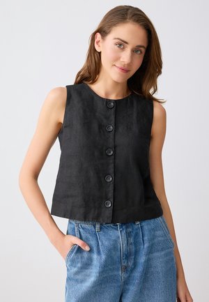 Jonge vrouw met lang bruin haar, gekleed in een mouwloze zwarte top met knopen en een hooggesneden blauwe spijkerbroek, met één hand in de zak.