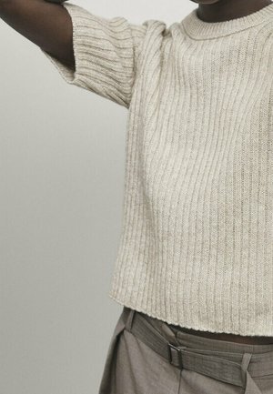 Jasnobeżowy prążkowany sweter z krótkim rękawem, noszony z szarymi spodniami z wysokim stanem i dopasowanym paskiem z tego samego materiału, na osobie o ciemnej karnacji.