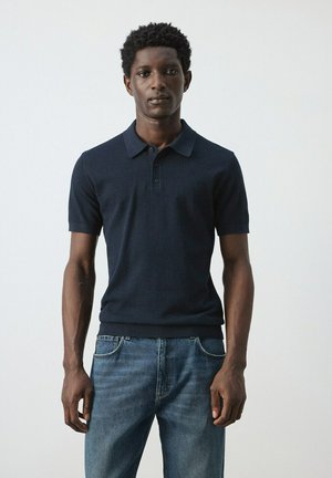 Hombre joven negro de pie, vistiendo una camiseta polo azul marino de manga corta y jeans azules contra un fondo claro y liso.