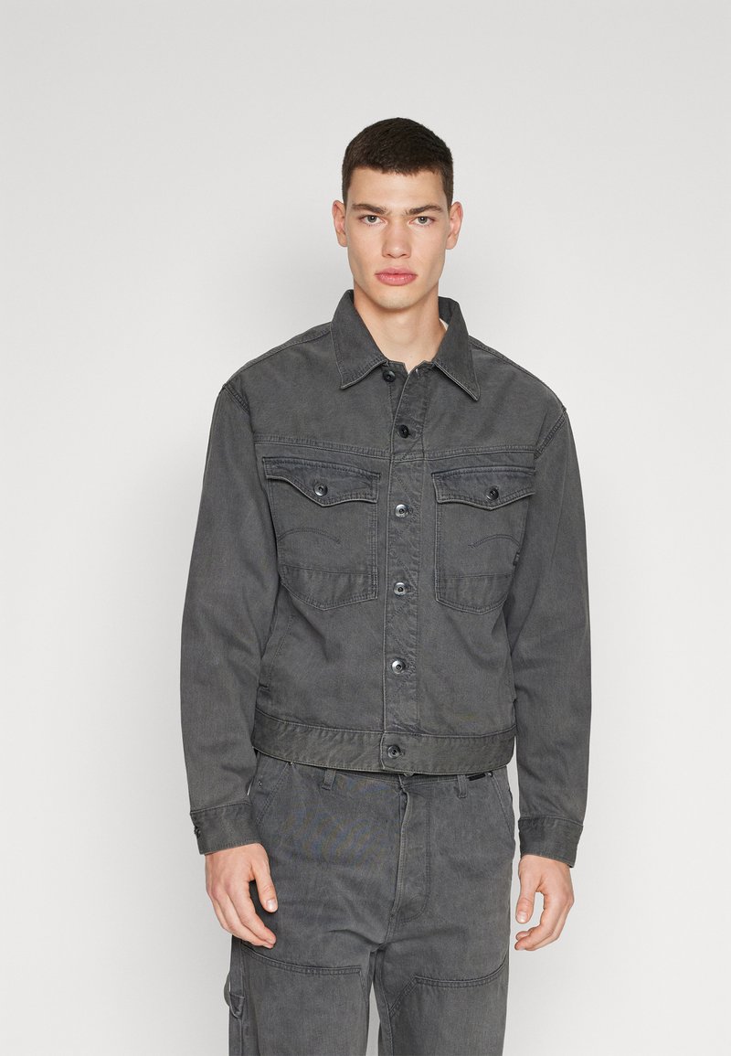 Γκρι τζάκετ από τζιν με γιακα, μπροστινό κούμπωμα με κουμπιά, δύο τσέπες στο στήθος και ορατές ραφές. Υφασμάτινη υφή με ελαφριά γυαλάδα.