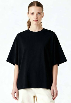 Jeune femme à la peau claire et aux cheveux bruns attachés en queue de cheval, portant une chemise noire ample à manches courtes et un pantalon clair, debout devant un fond blanc.