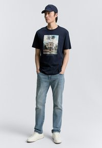 Tumši zili T-krekls ar grafisku attēlu, kurā attēlots ēka un palmu koki, savienots ar gaiši ziliem džinsiem un baltām kedām, kā arī ar tumši zilu cepuri.