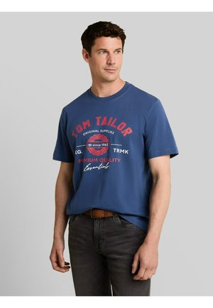 Man die een blauw Tom Tailor T-shirt draagt met rode en witte tekst, donkere spijkerbroek en een bruine riem, kijkt iets opzij.