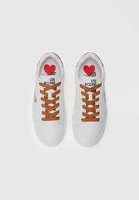 Bijele tenisice s smešnim vezicama, glatkim materijalom i ispisom "LOVE MOSCHINO" na jeziku, uz crvenu srčanu grafiku.