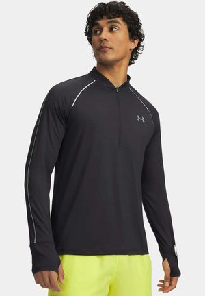 Mann trägt ein schwarzes langärmeliges Sportoberteil mit Reißverschluss, reflektierendem Besatz und Daumenlöchern, kombiniert mit leuchtend gelben Shorts.