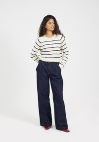 Donna che indossa un maglione bianco a righe navy, jeans larghi scuri e scarpe rosse, in piedi con le mani nelle tasche su uno sfondo semplice.