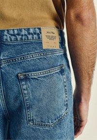 Modri kavbojki iz jeansa z teksturiranim izgledom. Opremljene s prometnim rjavim usnjenim oznako, ki vsebuje blagovno znamko in navodila za nego na hrbtni strani pasu.