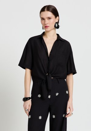 Femme portant une chemise noire à manches courtes boutonnée, nouée à la taille, un pantalon noir avec des motifs de fleurs blanches, un bracelet noir et de grandes boucles d'oreilles.