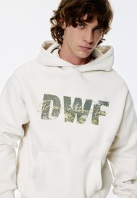Crèmekleurige hoodie met een groot, verweerd groen "DWF"-logo en cursieve tekst. Voorzien van een voorvak en een ruime capuchon.