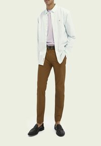 Hellblau gestreiftes Langarmhemd über einem fliederfarbenen T-Shirt, kombiniert mit braunen, schlank geschnittenen Hosen und schwarzen, strukturierten Loafers.