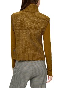 Khakifarbenes ärmelloses Rollkragenoberteil mit gerippter Textur, kombiniert mit einem langärmeligen Senfgelben Oberteil und karierter Hose.