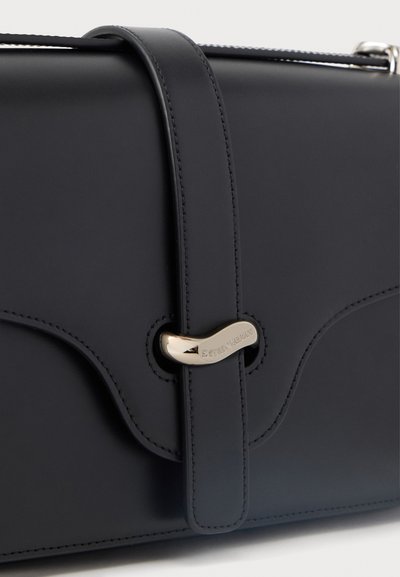 Μαύρη δερμάτινη τσάντα χειρός με καμπυλωτό καπάκι που ασφαλίζει με πόρπη σε χρυσή απόχρωση, χαραγμένη με το όνομα της μάρκας.