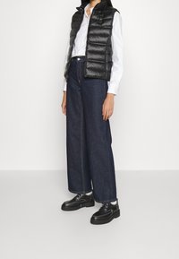 Czarna pikowana kamizelka z poziomym przeszyciem, zestawiona z białą koszulą i szerokimi ciemnymi dżinsami o kroju szerokich nogawek. Obuwie: czarne sznurowane buty.