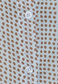 Lyseblå skjorte stof med et brunt pletmønster, med en klar plastknap og synlige sting langs knaplæsten.