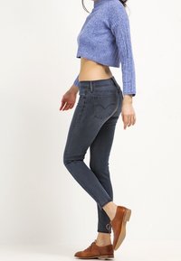 Jeans ajustados de color azul oscuro con bolsillos traseros, combinados con un suéter corto de textura azul claro y zapatos marrones con cordones.