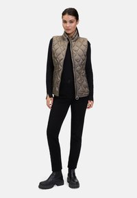 Beige Steppweste mit hohem Kragen, Reißverschluss und Seitentaschen, kombiniert mit einem schwarzen Langarmtop und schwarzen Hosen, ergänzt durch schwarze Stiefel.