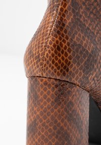 Chaussure à talons hauts marron avec une texture brillante en peau de serpent, talon bloc robuste et couture détaillée le long des coutures.