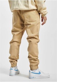 Beige joggingbroek met elastische boorden, voorzien van achterzakken. Gecombineerd met witte sneakers met blauwe accenten. Gladde textuur, casual pasvorm.
