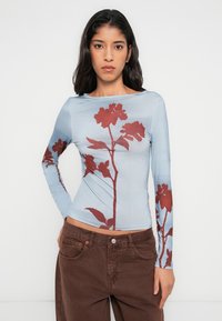 Μπλούζα με μακριά μανίκια σε ανοιχτό μπλε ύφασμα με κόκκινο σχέδιο λουλουδιών. Το υλικό φαίνεται λείο με εφαρμοστή γραμμή.