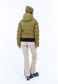 Olivgrüne Pufferjacke mit Kapuze, verkürztem Schnitt und geripptem schwarzen Saum, kombiniert mit beigen Hosen und schwarzen Skischuhen mit Reißverschlüssen.