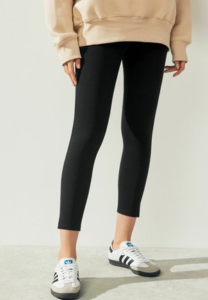 Personne portant des leggings noirs, un sweat beige et des baskets blanches Adidas avec des bandes noires, debout sur un sol clair contre un mur blanc.
